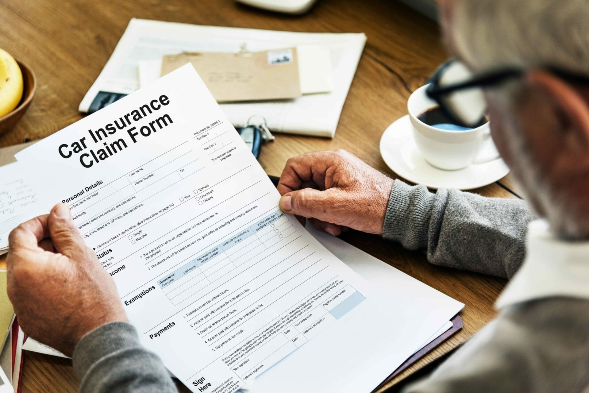 Man is reviewing a car insurance claim form at a desk with a cup of coffee, envelopes, and paperwork in the background.