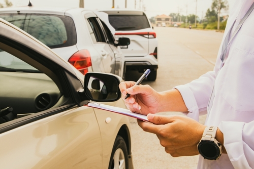 Person in a white coat writing on a clipboard while inspecting a car involved in a roadside incident.