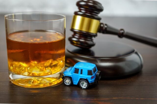 A judge's gavel on a courtroom table alongside a glass of whiskey and a miniature car, symbolizing the "no drinking and driving" concept.