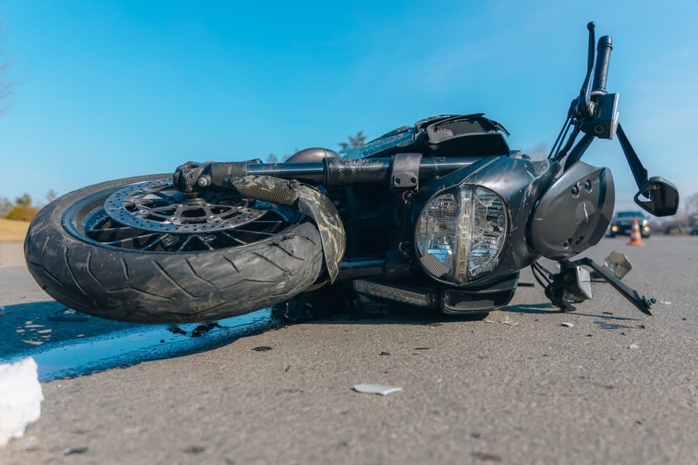 A black motorcycle lies sprawled on the asphalt, bearing the scars of a severe accident in a stark close-up. The scene speaks of lost control and a sudden fall.