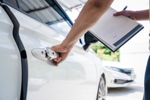 Person inspecting a white car and taking notes on a clipboard during an auto damage assessment.