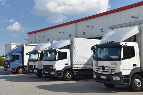 Four commercial delivery trucks parked in a row at a warehouse loading dock under a clear blue sky.