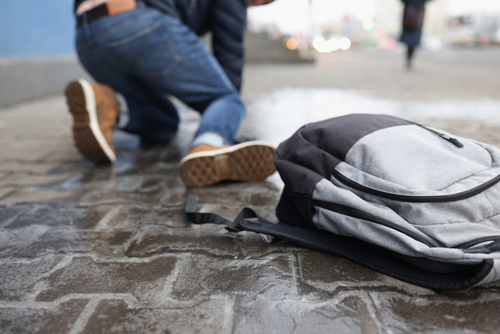 Person kneeling on icy pavement after a slip and fall, with a grey and black backpack on the ground nearby.