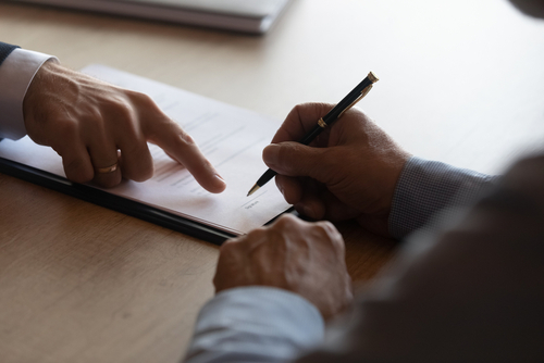 A person signing a legal document with a black pen while another individual points to a section on the paper, suggesting a contract review or agreement signing.