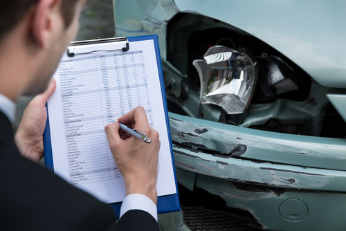 A claims adjuster fills out a vehicle damage report on a clipboard in front of a light blue car with a smashed headlight and dented bumper.