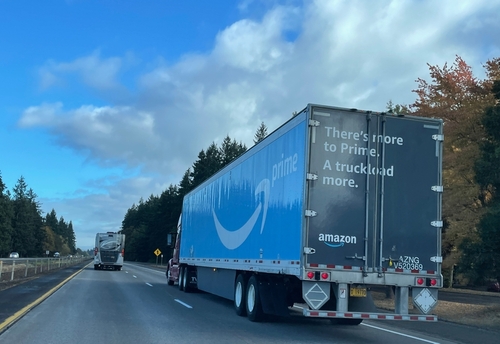 Amazon Prime semi-truck driving on a highway surrounded by trees under a partly cloudy sky, with the slogan "There's more to Prime. A truckload more." printed on the back of the trailer.