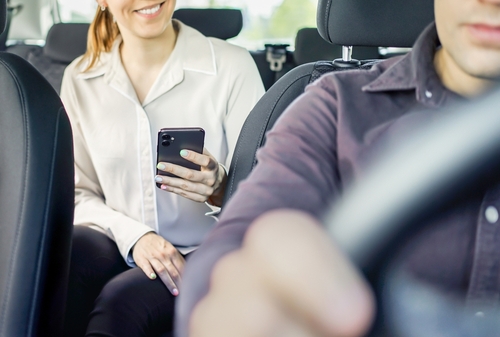 A smiling woman sits in the backseat of a car holding a smartphone, while the driver focuses on the road ahead.