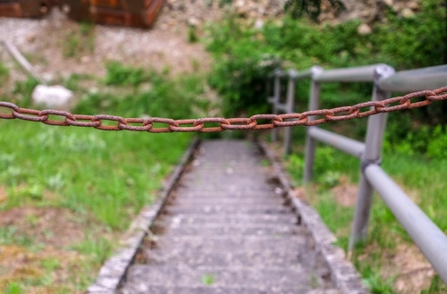 A rusted chain blocks access to a steep outdoor staircase with metal handrails, surrounded by grass and trees, indicating a potential safety hazard.