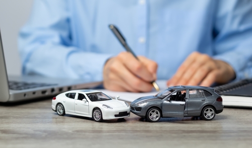 Two toy cars representing a car accident sit on a desk in front of a person writing, with a laptop nearby in a legal or insurance setting.