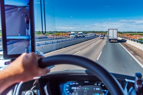 View from inside a commercial truck showing the driver's hand on the steering wheel while driving on a multi-lane highway with clear skies and light traffic.