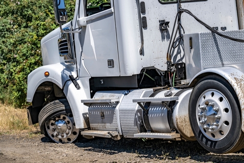 Close-up of a damaged white semi-truck with a flat front tire, parked on a dirt roadside near green foliage.