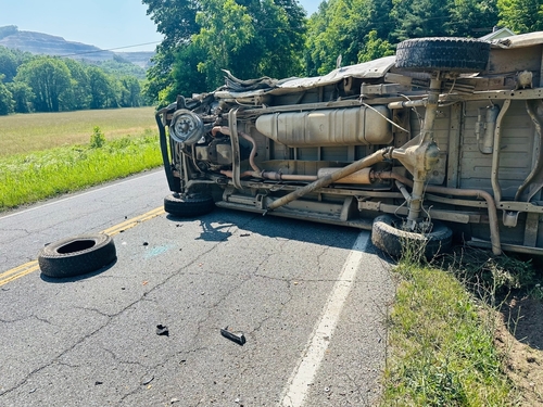 An overturned truck lying on its side on a two-lane rural road, with a detached wheel and debris scattered on the pavement, surrounded by green trees and a grassy field.