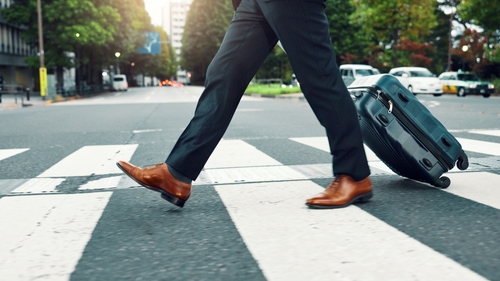 Person pulling a suitcase while walking across a pedestrian crosswalk in an urban setting, with cars and trees in the background.