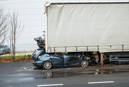 A severely damaged black car wedged under the rear of a large white semi-truck trailer after a collision on a wet road near an industrial building.